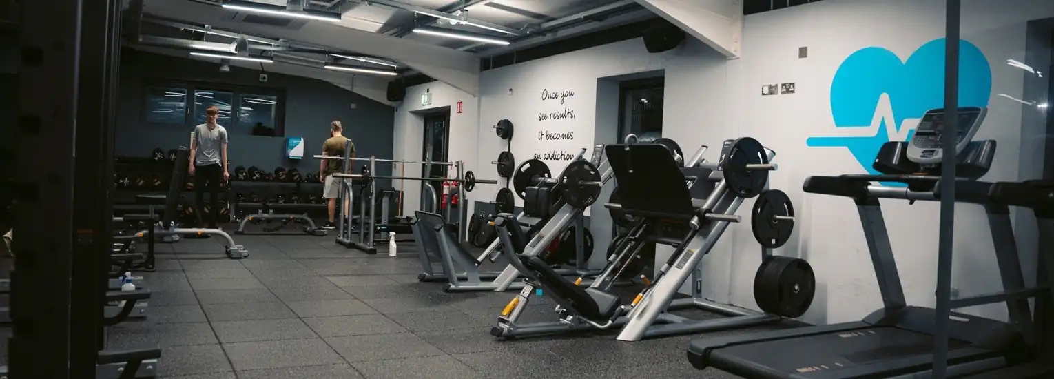 A modern gym with various exercise machines, weights, and benches. Two people are standing near the back wall, which has motivational writing and a blue heart graphic. The gym is well-lit and spacious.