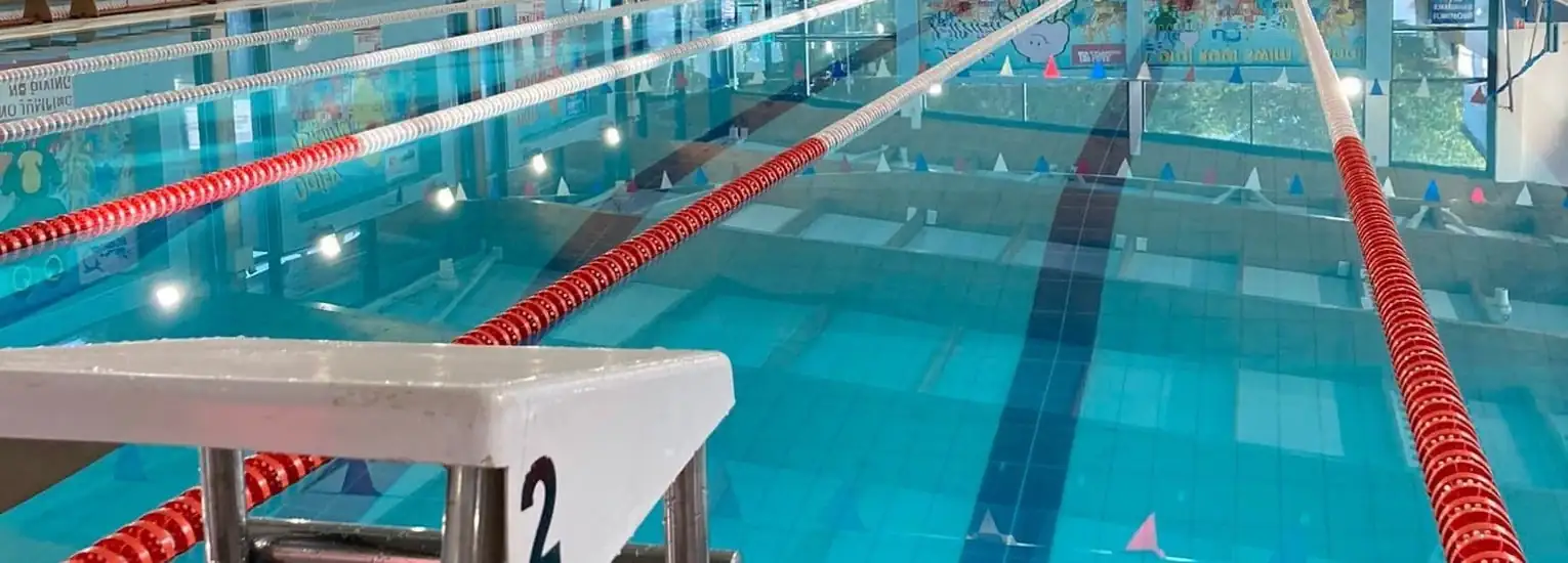 Close-up view of a swimming pool with clearly marked lanes, red lane dividers, and a starting block labelled “2” in the foreground. Colourful wall decorations are visible in the background.