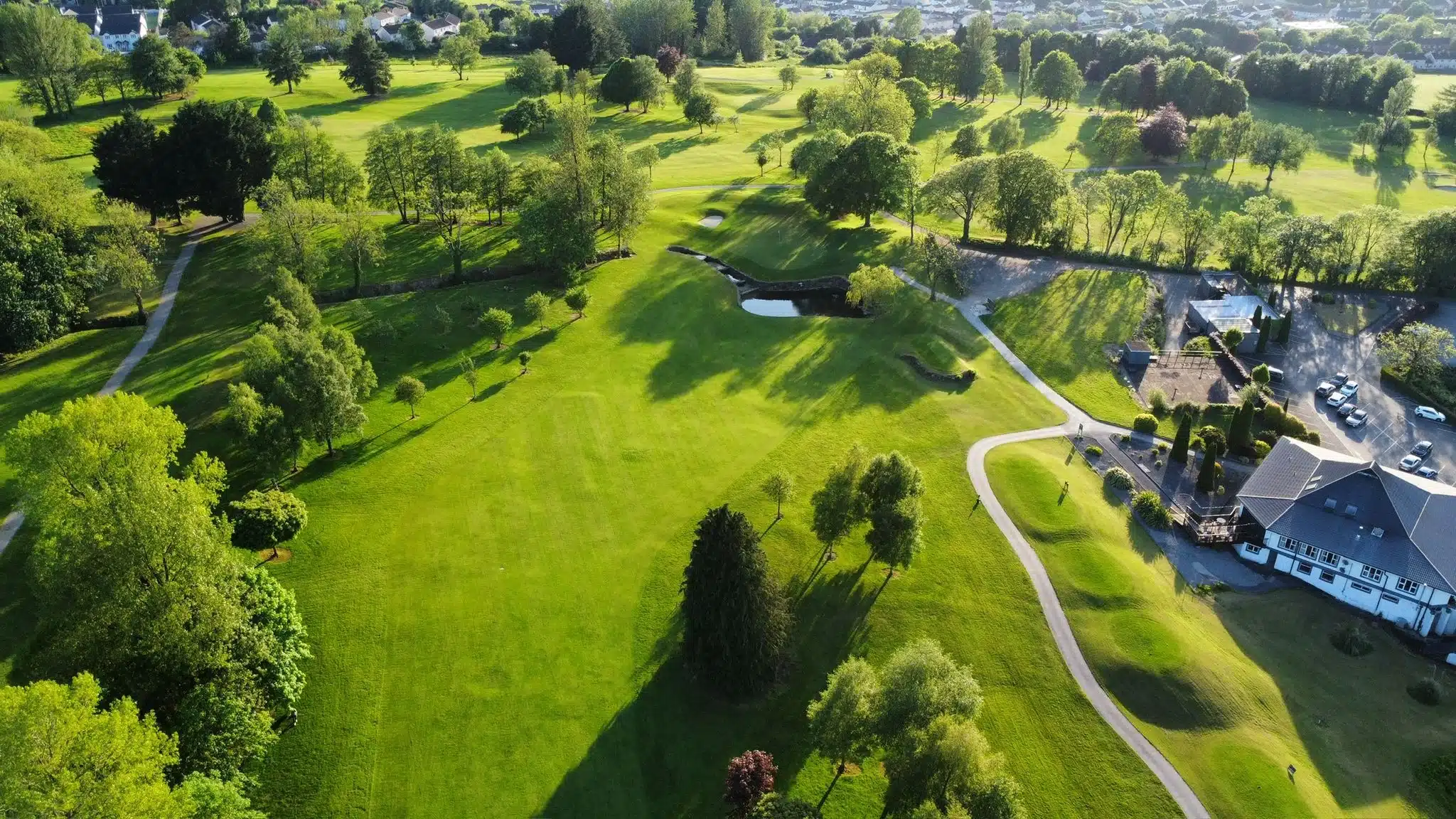 Aerial view of a lush green golf course with winding paths, sand bunkers, scattered trees, a clubhouse, and parked cars on a sunny day.