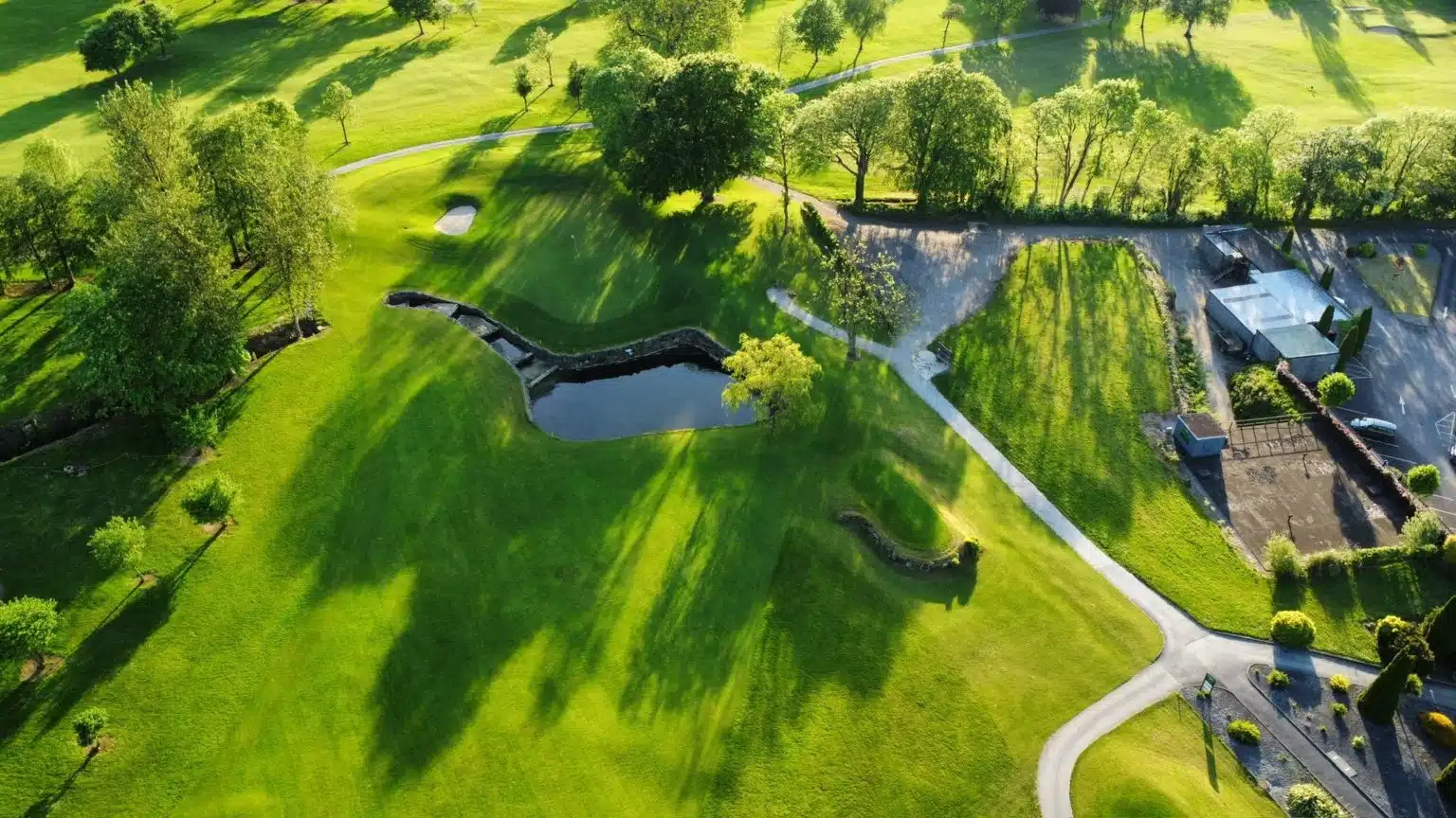 Aerial view of a green golf course with trees casting long shadows, a small pond, winding pathways, and manicured lawns on a sunny day.