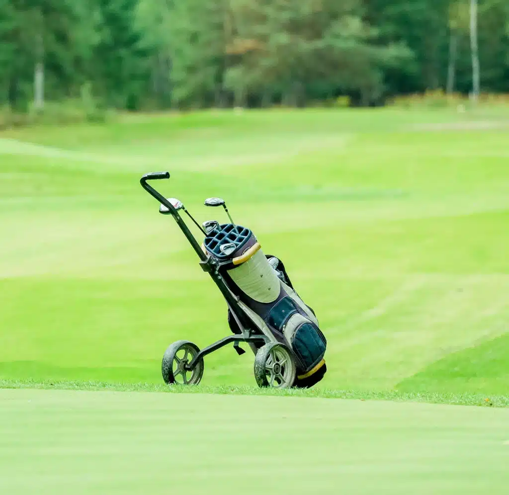 A golf push trolley with a golf bag stands on a grassy golf course, with trees and green fairways in the background.