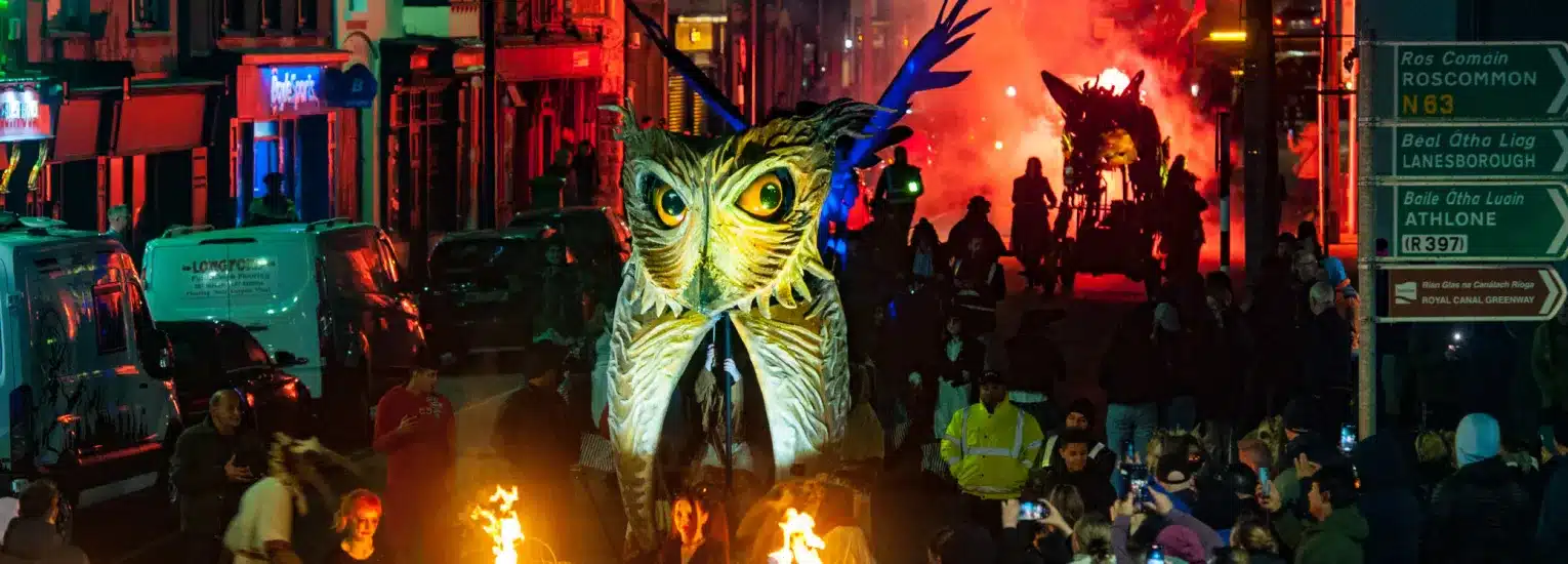 A large illuminated owl puppet is paraded through a crowded, colourful city street at night, with people watching and taking photos. Fire performers and another glowing creature are visible further down the street.