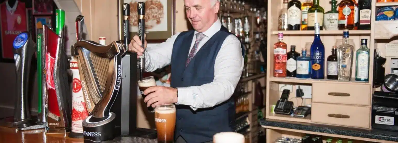 A barman in a waistcoat and tie pours a pint of Guinness behind a well-stocked bar with bottles of spirits, beer taps, and a till.