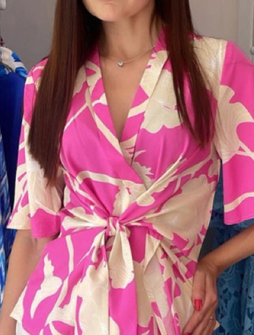 Woman wearing a pink and cream floral wrap dress with a front knot, standing with one hand on her hip. Only her upper body is visible; her face is partially out of shot.