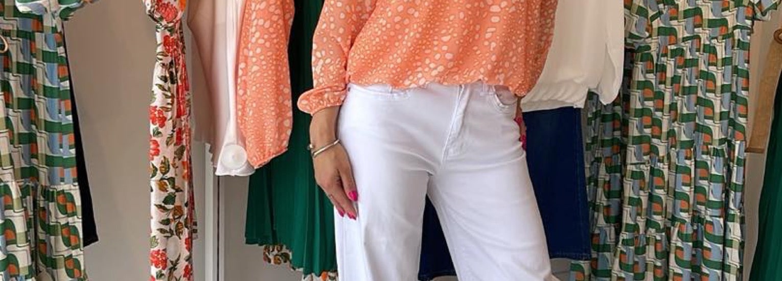 A person stands indoors wearing a peach-patterned blouse and white trousers, surrounded by colourful clothes hanging on a rail, including floral, green, and geometric patterned garments.