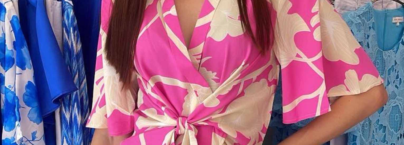 A woman wearing a pink and cream floral wrap dress poses with one hand on her hip in front of a rail of blue and white dresses.