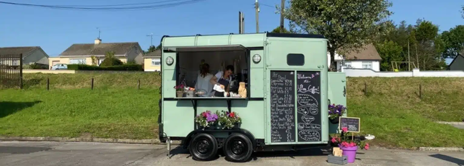 A mint green food truck with a large serving window and chalkboard menu is parked by a grassy area. Two people are inside serving customers. Potted flowers decorate the front of the lorry. Houses and trees are in the background.