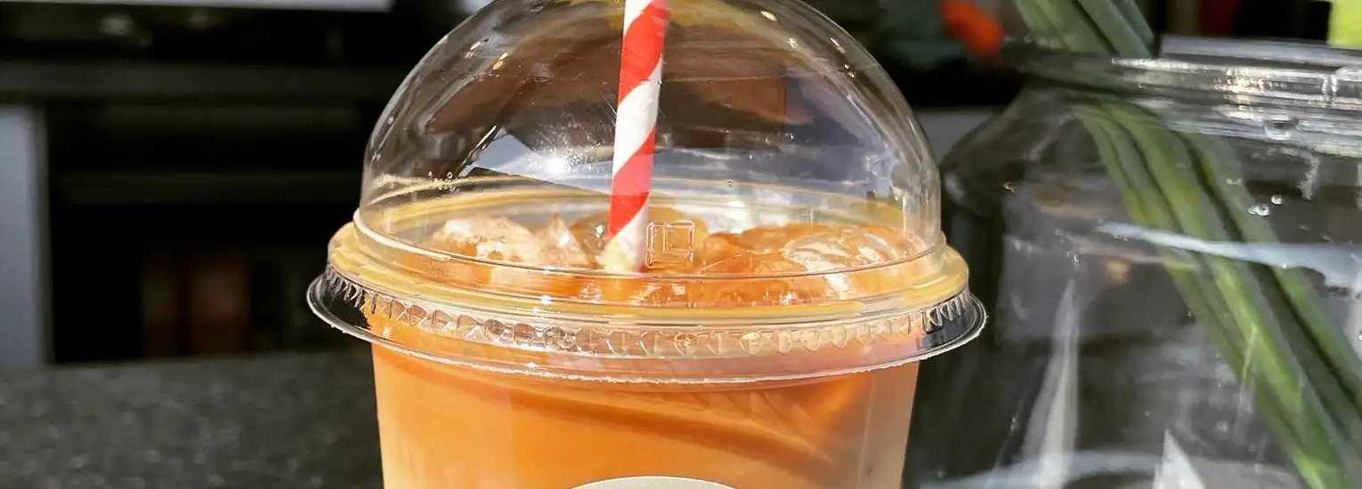 A plastic cup of iced coffee with a dome lid and a red-and-white striped straw sits on a counter next to a glass vase with green stems.