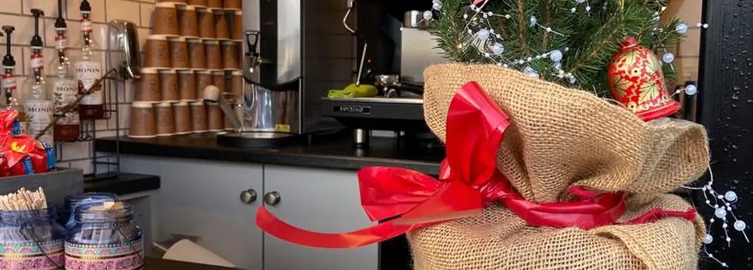 A small Christmas tree in a hessian sack with a red ribbon sits on a café counter, decorated with baubles and lights. Behind it, takeaway coffee cups and drink bottles are stacked near a coffee machine.