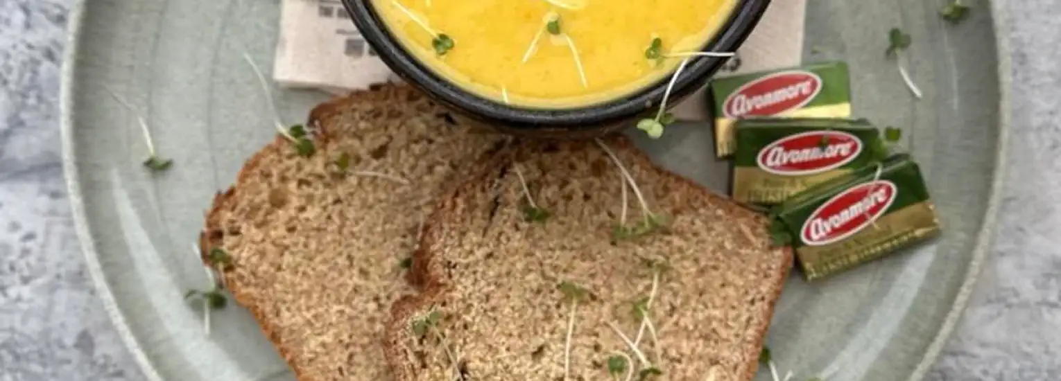 A plate with two slices of brown bread topped with sprouted seeds, a bowl of yellow soup, and three wrapped portions of butter.