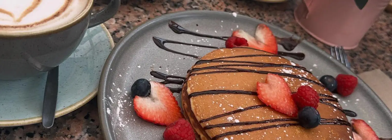 A plate of pancakes topped with sliced strawberries, blueberries, raspberries, icing sugar, and chocolate drizzle sits beside a cup of coffee with froth art on a marble tabletop.