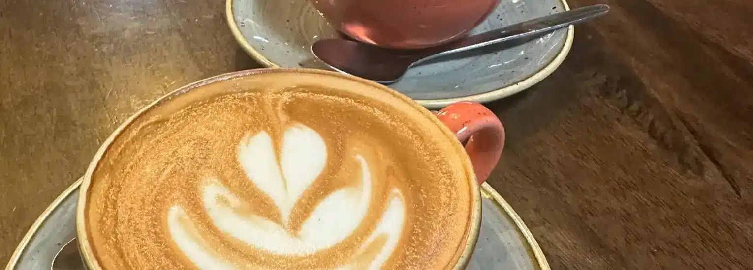 Two ceramic cups on saucers sit on a wooden table. The front cup contains a latte with a leaf-shaped foam design, and the other cup in the background has a spoon resting on its saucer.