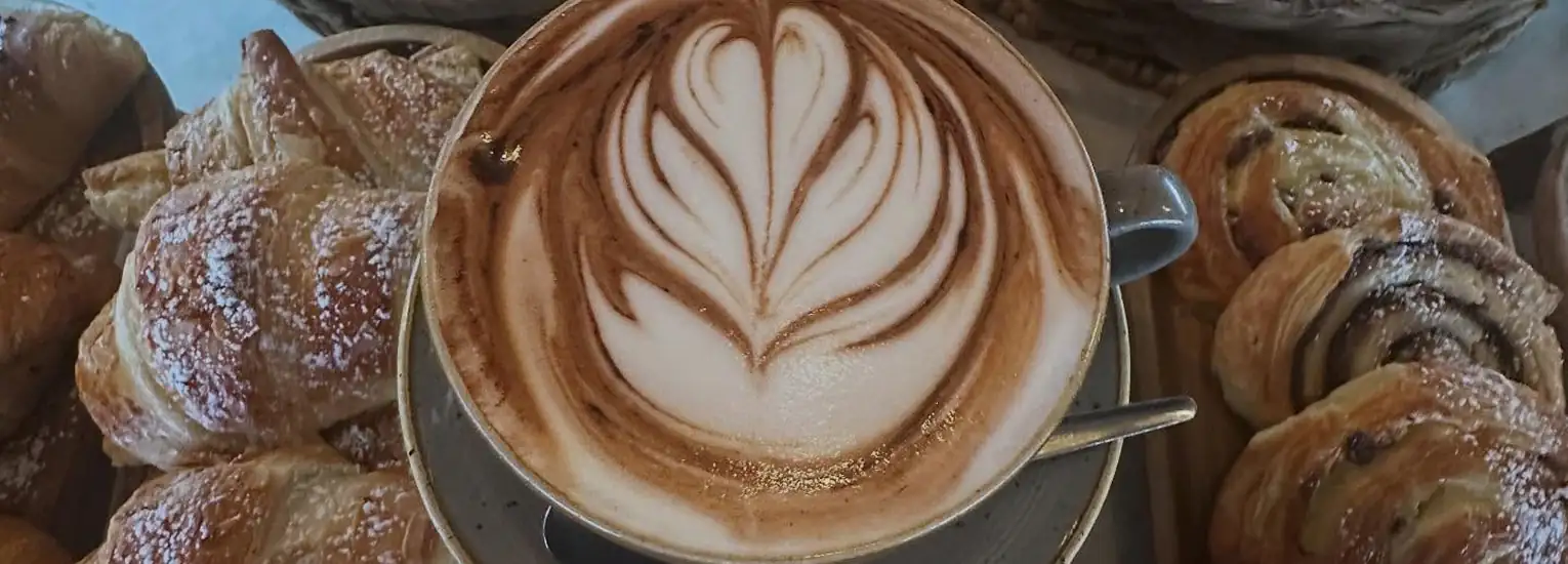 A cup of cappuccino with intricate latte art sits on a saucer, surrounded by croissants and pastries dusted with icing sugar.