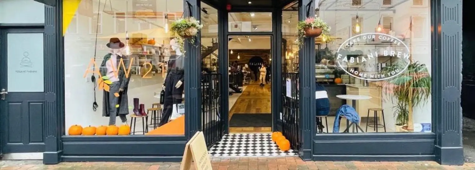 Shopfront with large glass windows displaying clothes and hats, pumpkins on the ground, and a sign above the door that reads "Abia Brew Home With You." The entrance has chequered tiles and hanging flower baskets.