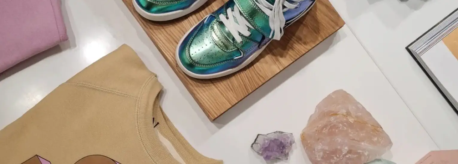 A display featuring a tan sweatshirt with a graphic, shiny green and blue trainers on a wooden board, several crystals, and an open book on a white surface.