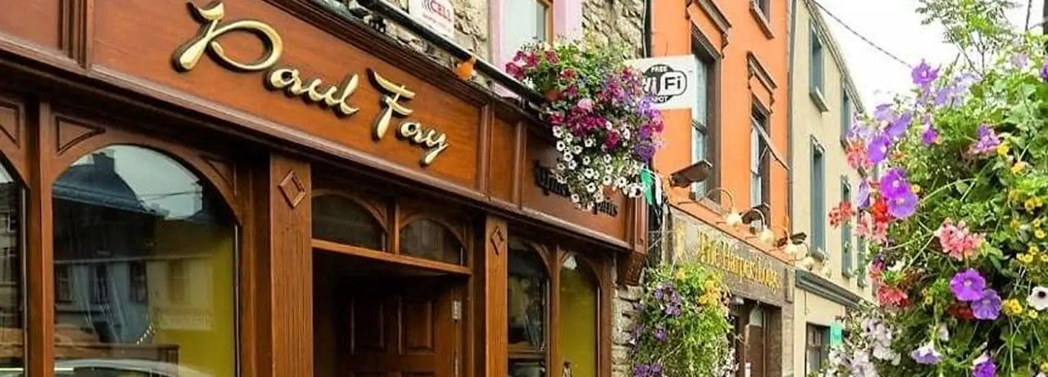 A charming street scene shows a wooden shopfront with the name "Paul Fay" in gold lettering, colourful hanging flower baskets, and vibrant buildings adorned with more flowers in the background.