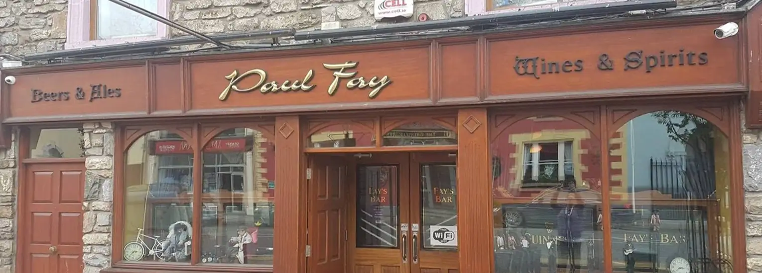 A traditional pub exterior with wooden frames and signage reading "Beers & Ales," "Paul Fay," and "Wines & Spirits." The entrance is open, and the stone building reflects a street scene in the windows.