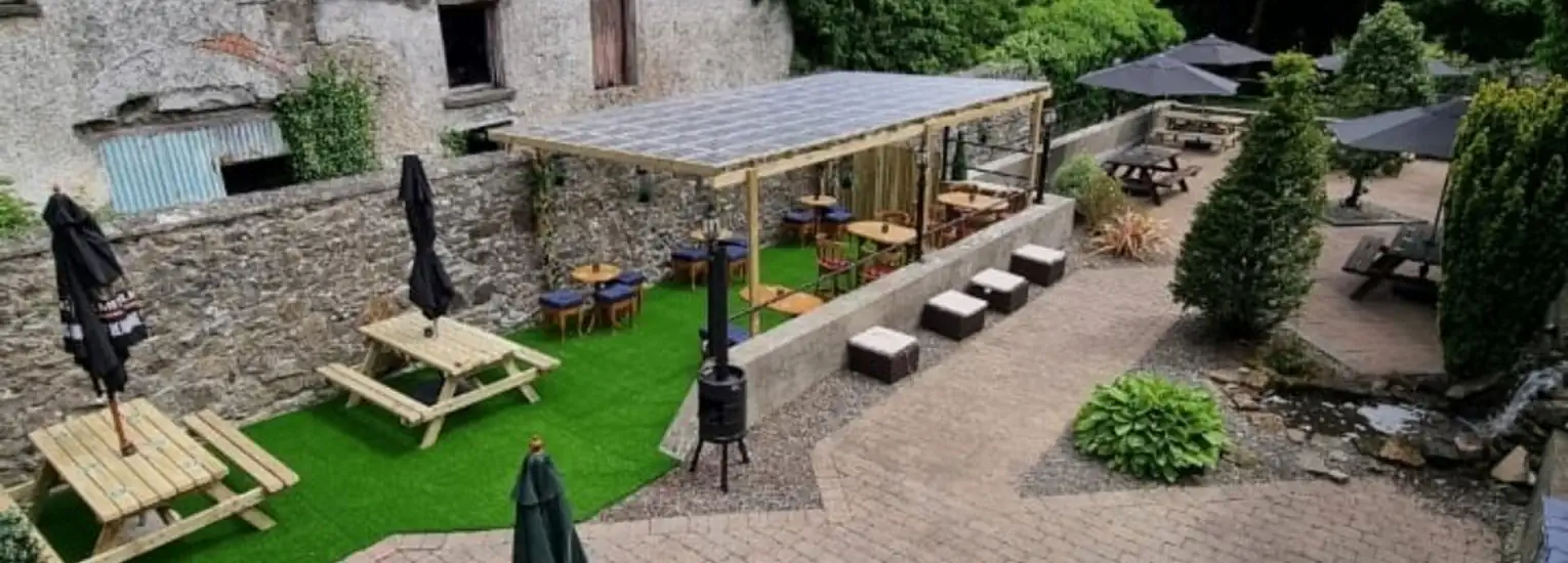 Outdoor seating area with wooden tables and benches, some under a pergola, others with umbrellas. Surrounded by stone walls, greenery, and a paved walkway, creating a cosy, rustic garden atmosphere.