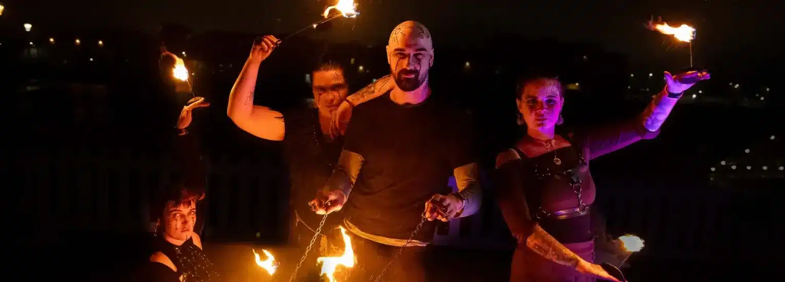 Four people perform with fire at night, holding flaming props and sticks, illuminated by the fire against a dark background. They wear dark clothing and dramatic expressions, creating a striking and intense scene.
