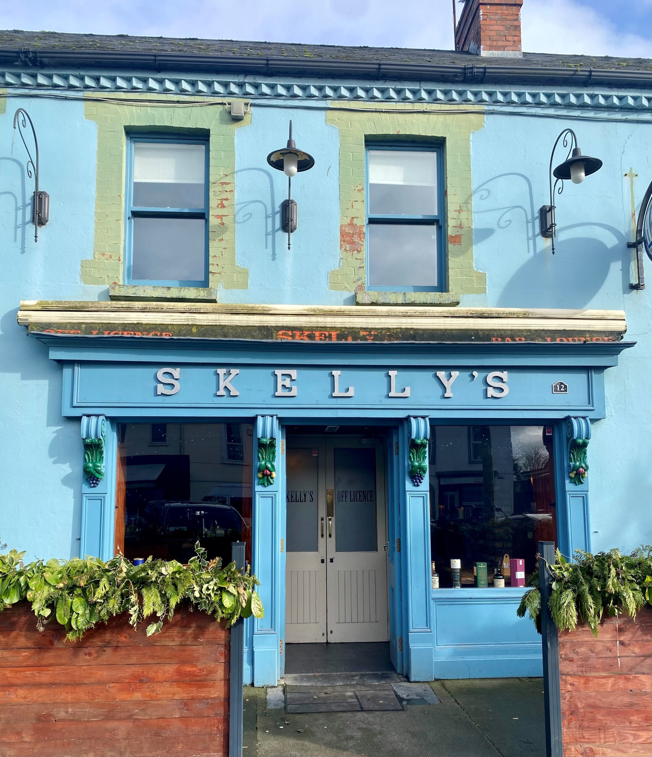 A blue-painted building with green trim, featuring a sign that reads "SKELLY'S" above double doors. There are outdoor lights, windows, and planters with greenery in front.