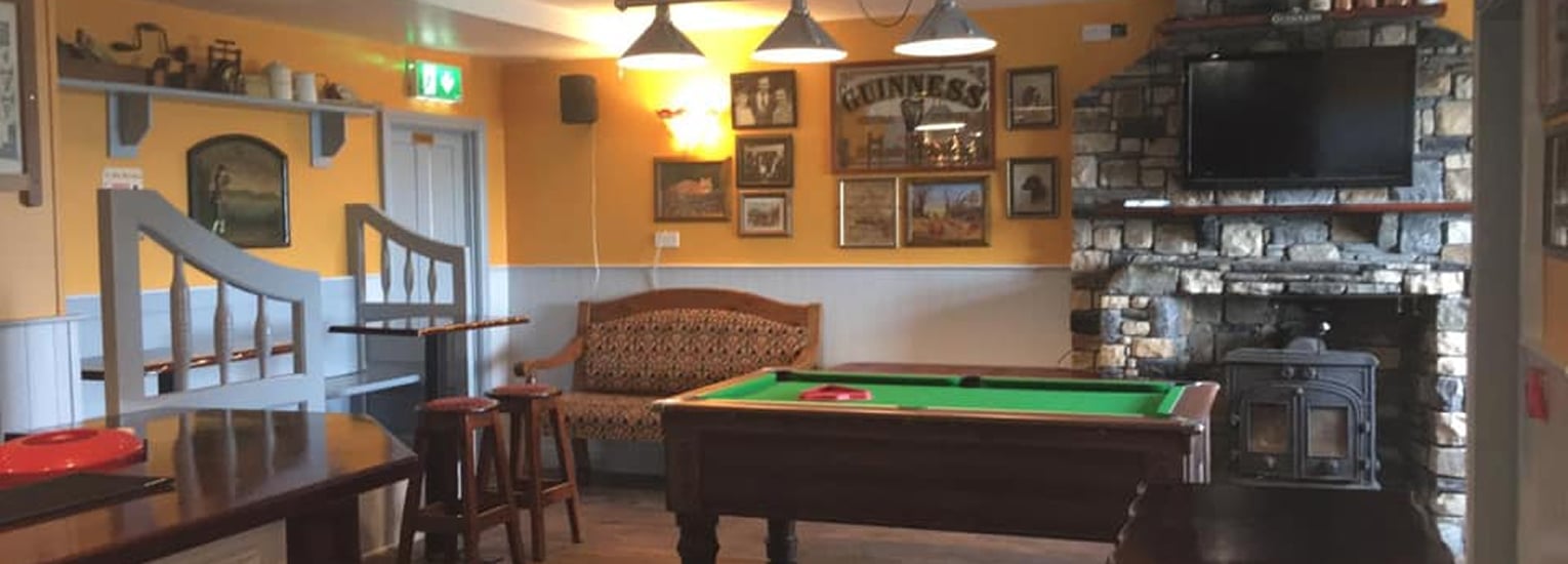 A cosy pub interior with a pool table, wooden furniture, a cushioned bench, framed pictures on yellow walls, and a stone fireplace beneath a mounted TV. Warm lighting hangs from the ceiling.