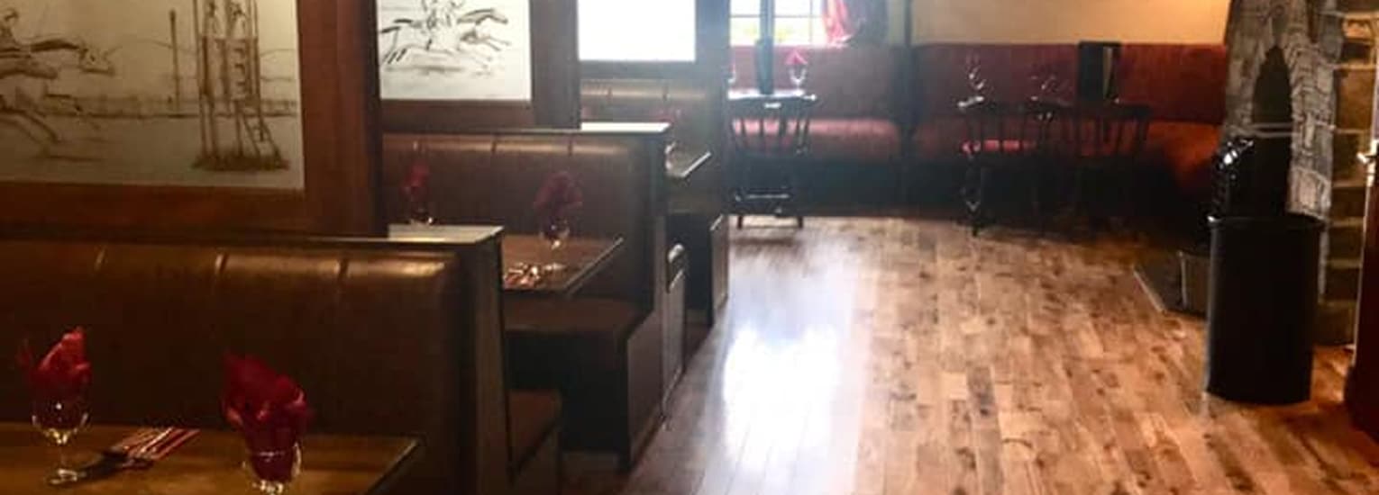 A cosy restaurant interior with wooden floors, brown booth seating on the left, dark wooden chairs and tables near a window, and soft natural light filling the space. Red serviettes are set on the tables.