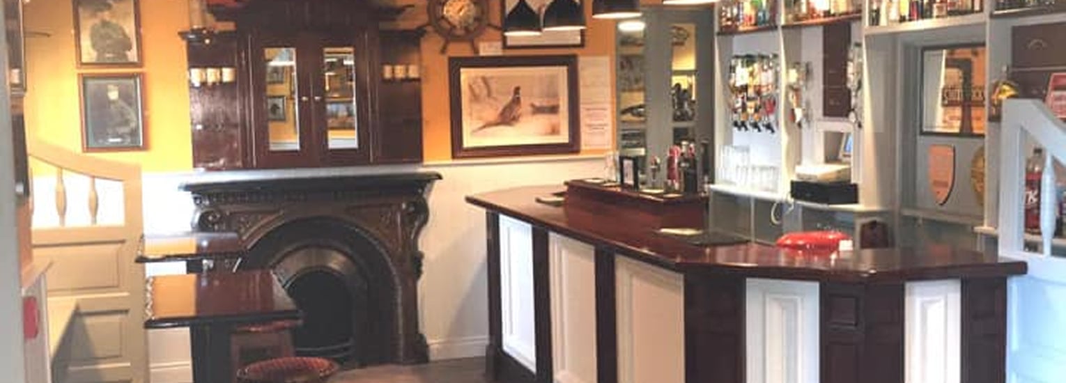 A cosy pub interior with a wooden bar counter, dark wood fireplace, wall-mounted shelves holding bottles, framed pictures, and small tables with chairs. Warm lighting creates a welcoming atmosphere.
