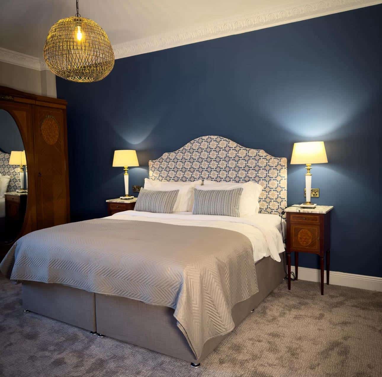 A stylish bedroom with a large bed, patterned headboard, beige bedding, two bedside tables with lamps, a wooden wardrobe with a mirror, blue feature wall, and a woven pendant light hanging from the ceiling.