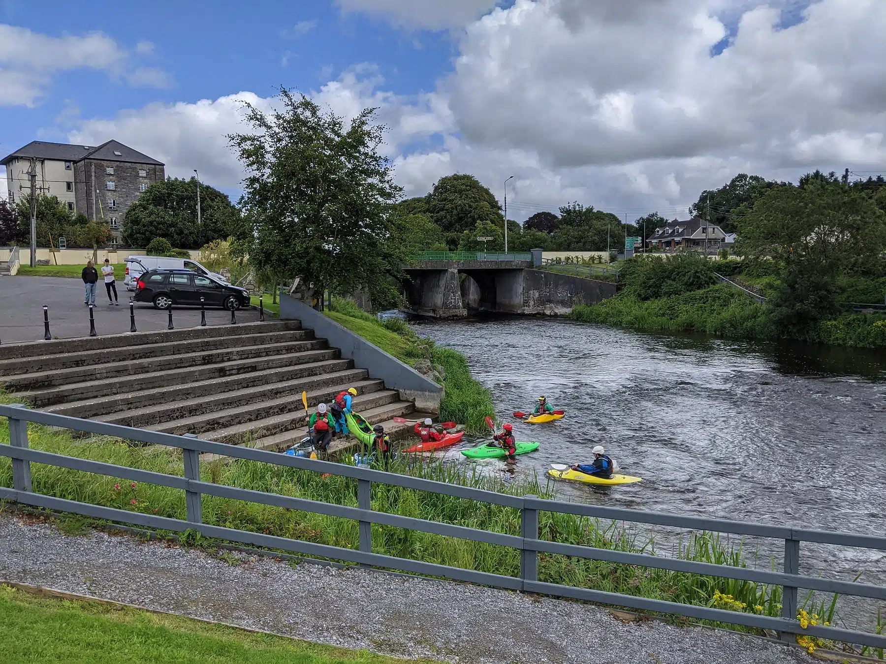 A group of people in colourful kayaks are gathered near the riverbank by a set of steps, with a bridge, grassy areas, and buildings in the background under a partly cloudy sky.