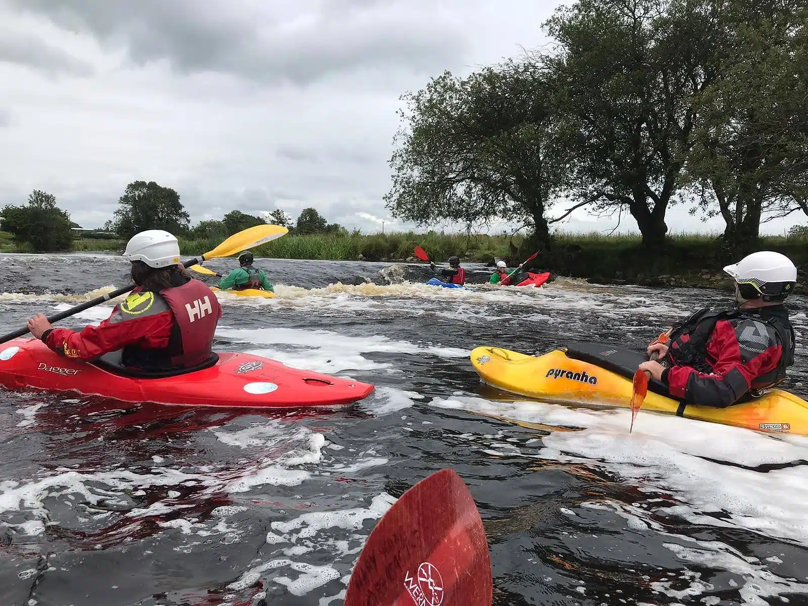 Several people in helmets and life jackets kayak on a river with foamy water under a cloudy sky, surrounded by trees and grassy banks.