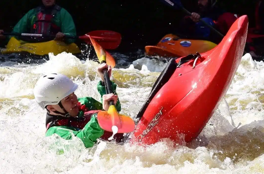 A person wearing a white helmet and green jacket is kayaking in rough water, tilting their red kayak whilst holding a paddle. Other kayakers are seen in the background, navigating the rapids.