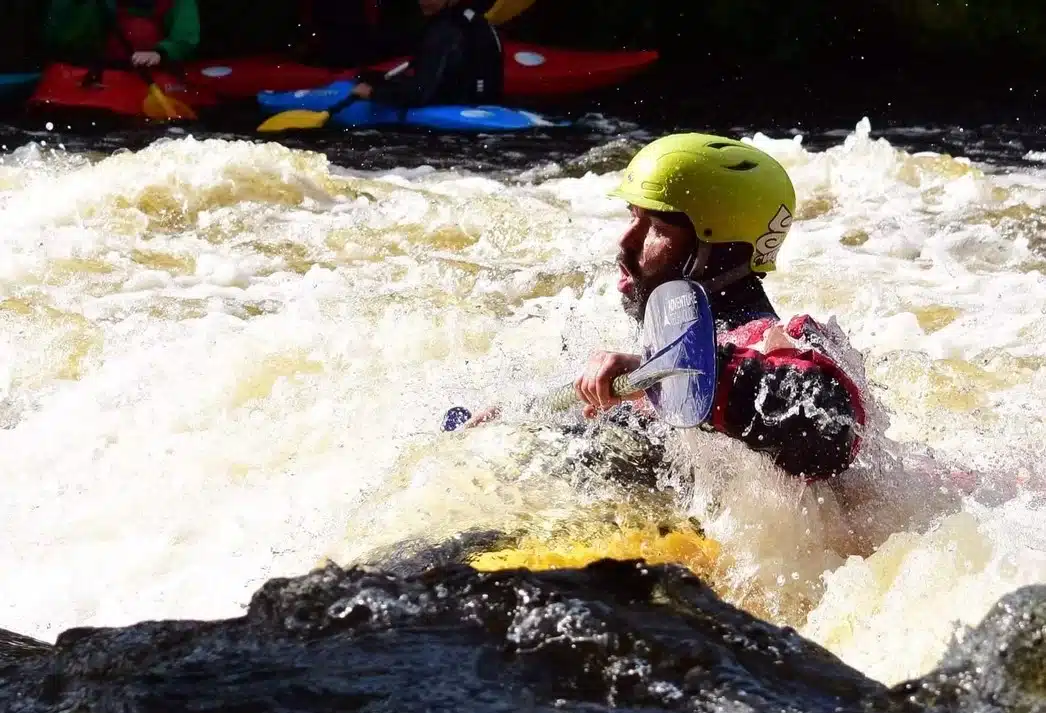 A person wearing a yellow helmet and life vest kayaks through rough, white-water rapids. In the background, other kayakers in colourful kayaks paddle on the river.