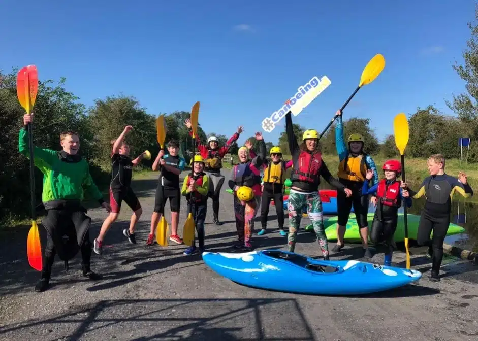A group of people in wetsuits and helmets stand outdoors holding paddles, some posing energetically beside a blue kayak. One person holds a sign that says "#Canoeing." The sky is clear and sunny.