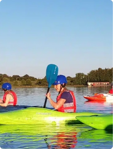 A person wearing a blue helmet and red life jacket kayaks on a calm lake, holding a blue paddle. Other kayakers are visible in the background under a clear sky.
