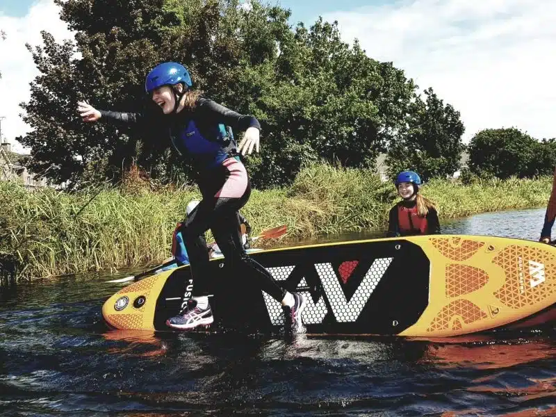 A person in a wetsuit and helmet excitedly jumps off a large paddleboard into the water, whilst another person in similar gear sits on the board, surrounded by reeds and trees.