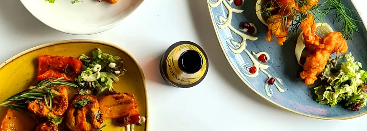 Three colourful plates of food, each with different vibrant dishes including grilled vegetables and garnishes, are arranged around a small bottle on a white table.