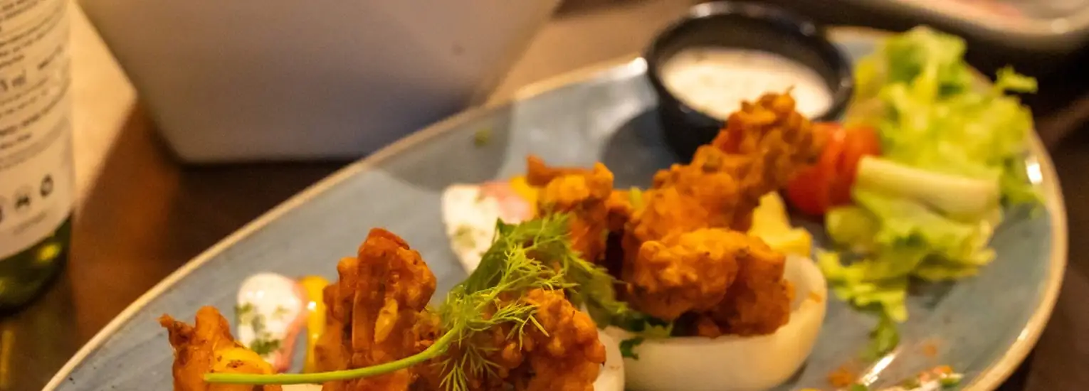 A close-up of a blue plate with crispy fried chicken pieces garnished with herbs, a side of dipping sauce, and a small salad with lettuce and cherry tomatoes.