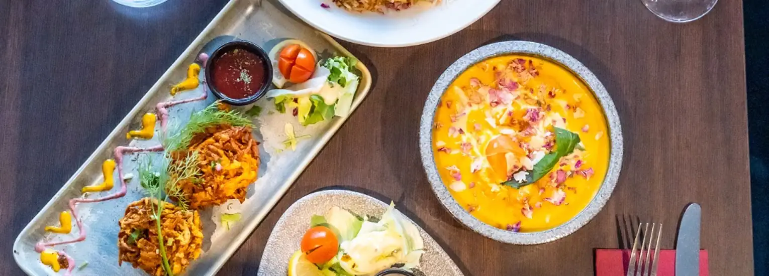 A table set with colourful Indian dishes, including a bright orange curry, vegetable fritters with sauce, garnished salads, and glasses of water, viewed from above. Plates and cutlery are arranged neatly on the dark wooden surface.