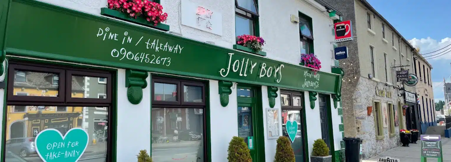 A green and white shopfront called "Jolly Boy" with flowers above the door, signs for eat-in and takeaway, and a heart-shaped sign in the window. Other businesses and cars line the street outside.
