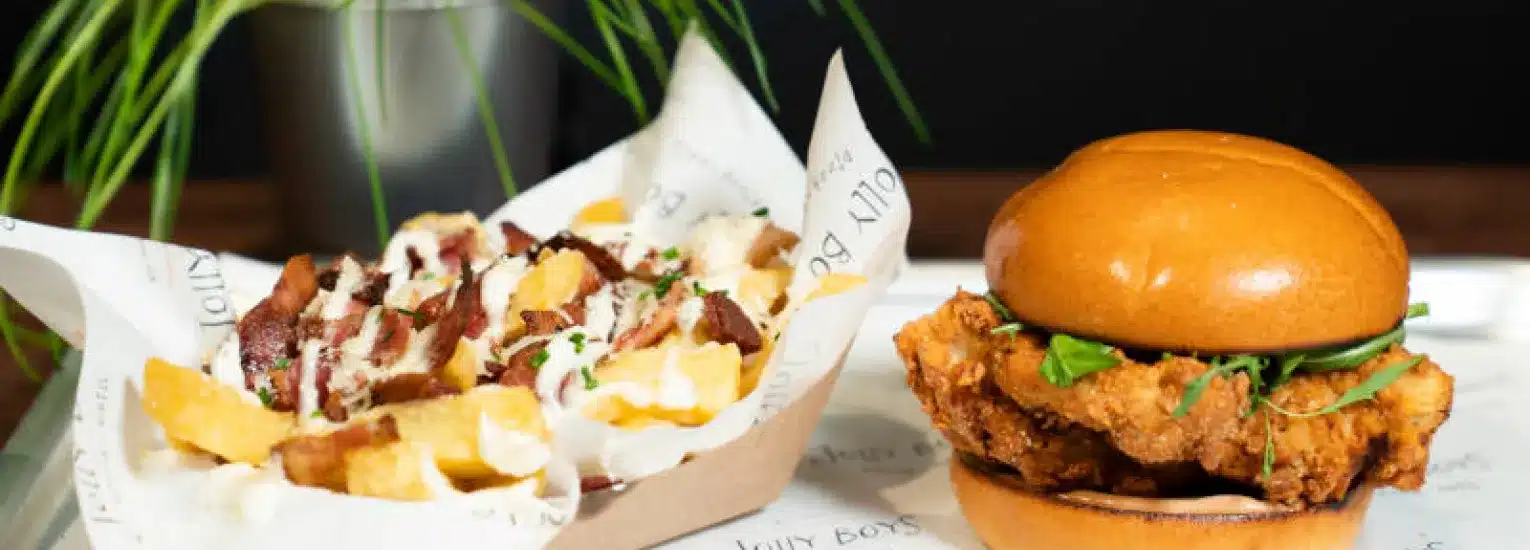 A tray with loaded chips topped with sauce and chopped meat in a paper container next to a fried chicken sandwich with greens in a shiny bun. A potted plant is blurred in the background.
