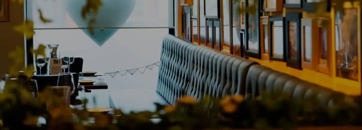 A cosy restaurant interior with a long, tufted bench, neatly set tables, framed pictures on the wall, and soft natural light filtering through a large window.