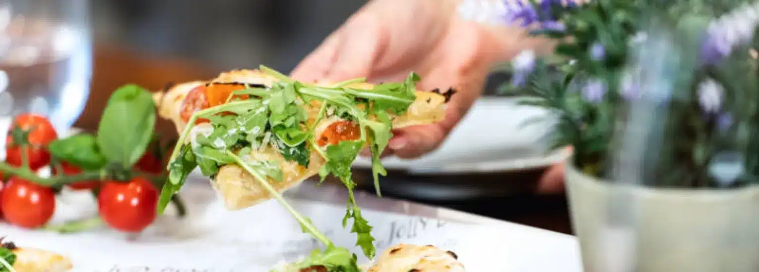A hand holds a slice of pizza topped with fresh rocket and cherry tomatoes, with more tomatoes and green herbs on the table nearby. A glass of water and potted plant are in the background.