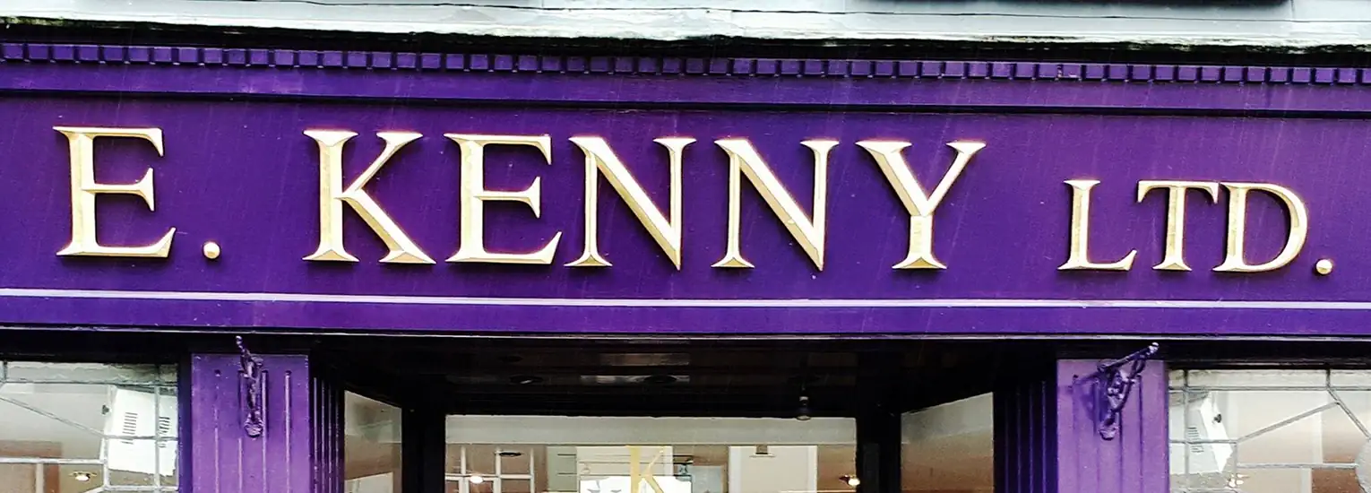 A shopfront sign with gold lettering on a purple background reads "E. KENNY LTD." The sign is above a glass door and window.