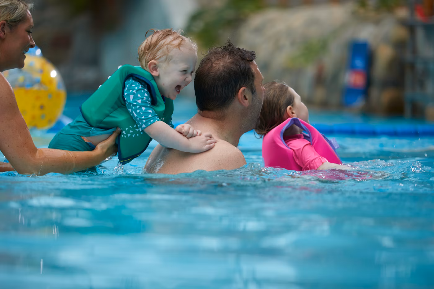 A smiling adult in a swimming pool holds a small child in a pink float, while another happy child wearing a green flotation vest is supported by a woman and plays nearby.