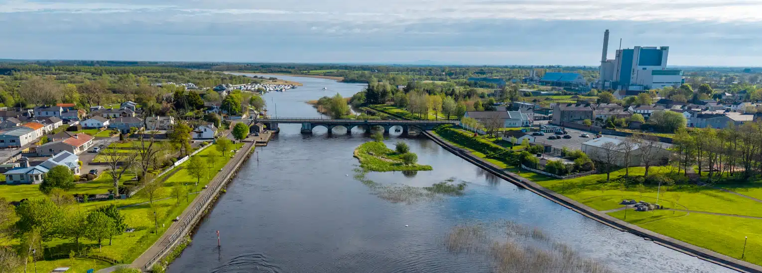 Aerial view of a river with a stone bridge, surrounded by green fields, houses, and trees, with a marina and a large industrial building visible in the background under a partly cloudy sky.