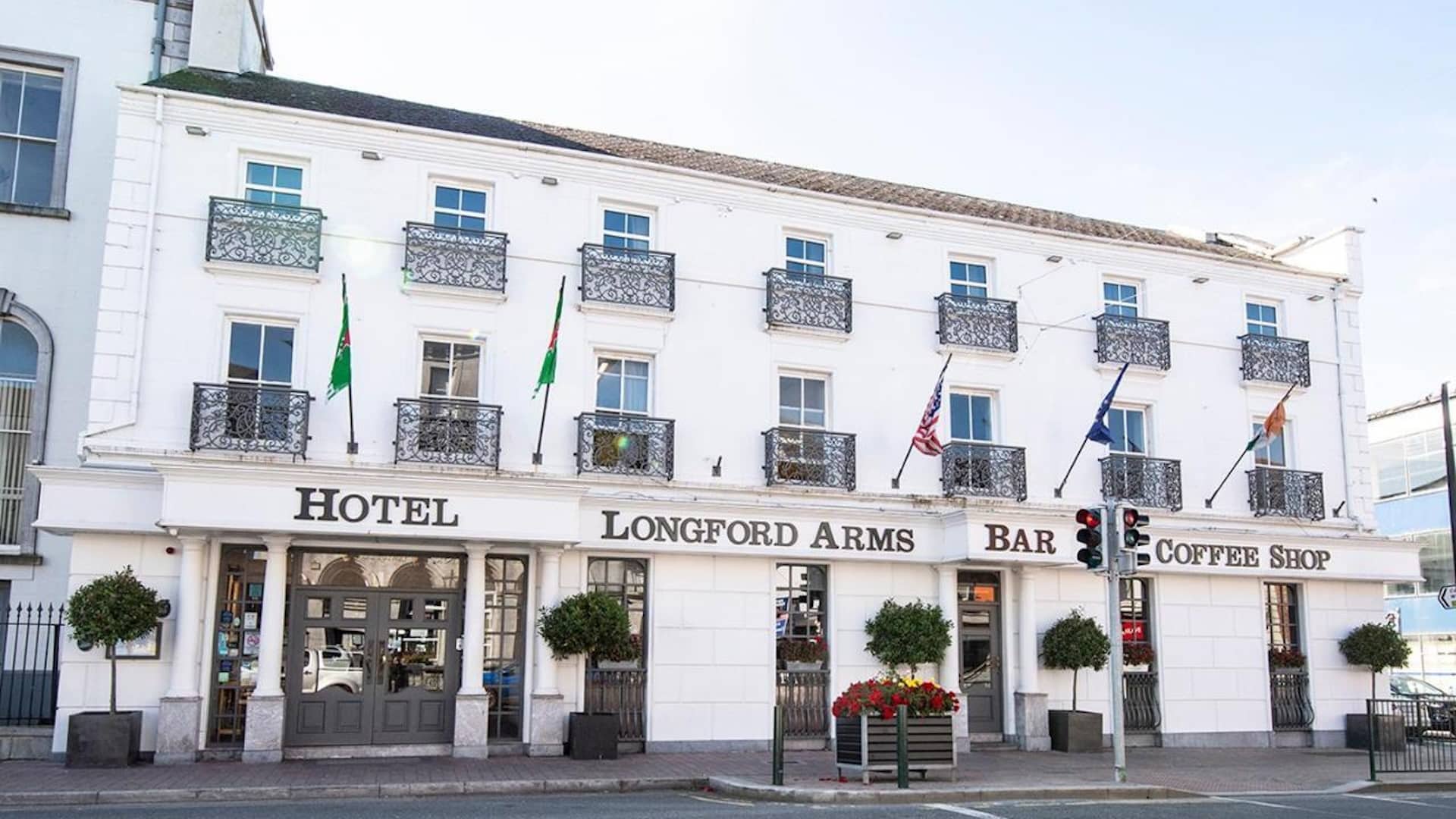 A white three-storey building with decorative balconies, flags, and signs reading "Hotel Longford Arms," "Bar," and "Coffee Shop." There are potted trees and flowers outside the entrance.