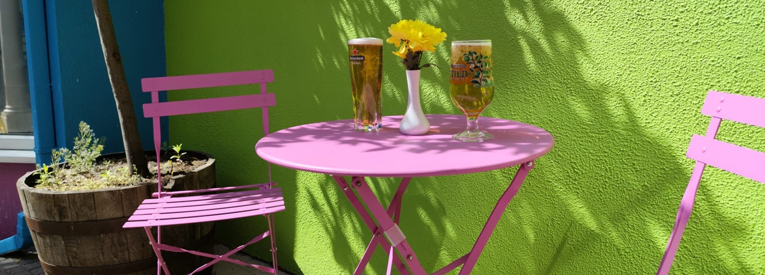 A pink metal table with two matching chairs, holding two pints of beer and a white vase with yellow flowers, sits against a bright green wall in an outdoor setting.