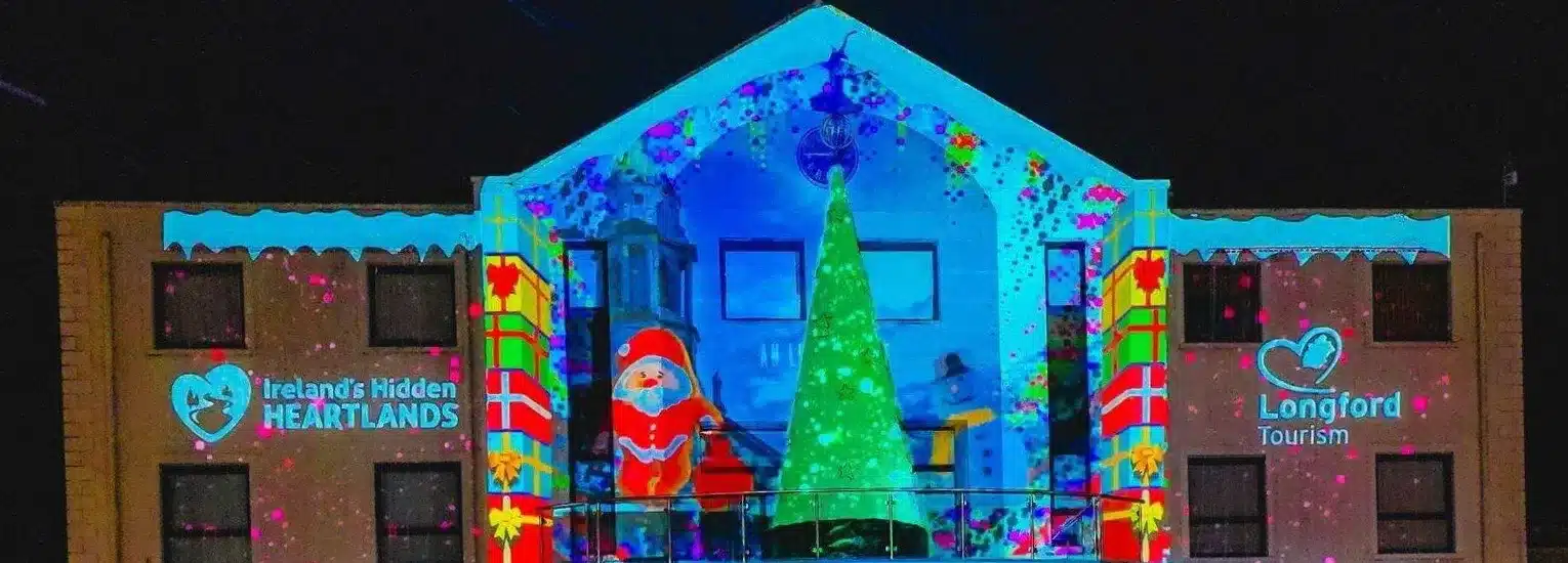 A building is lit up with colourful Christmas projections, including a large green Christmas tree, a Father Christmas figure, and festive decorations. Logos for