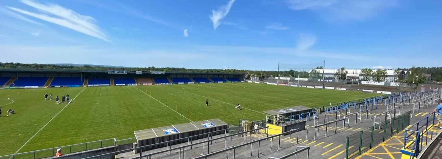 A panoramic view of an empty sports stadium with green grass, blue seating, and a few people practising on the pitch under a clear blue sky with light clouds.