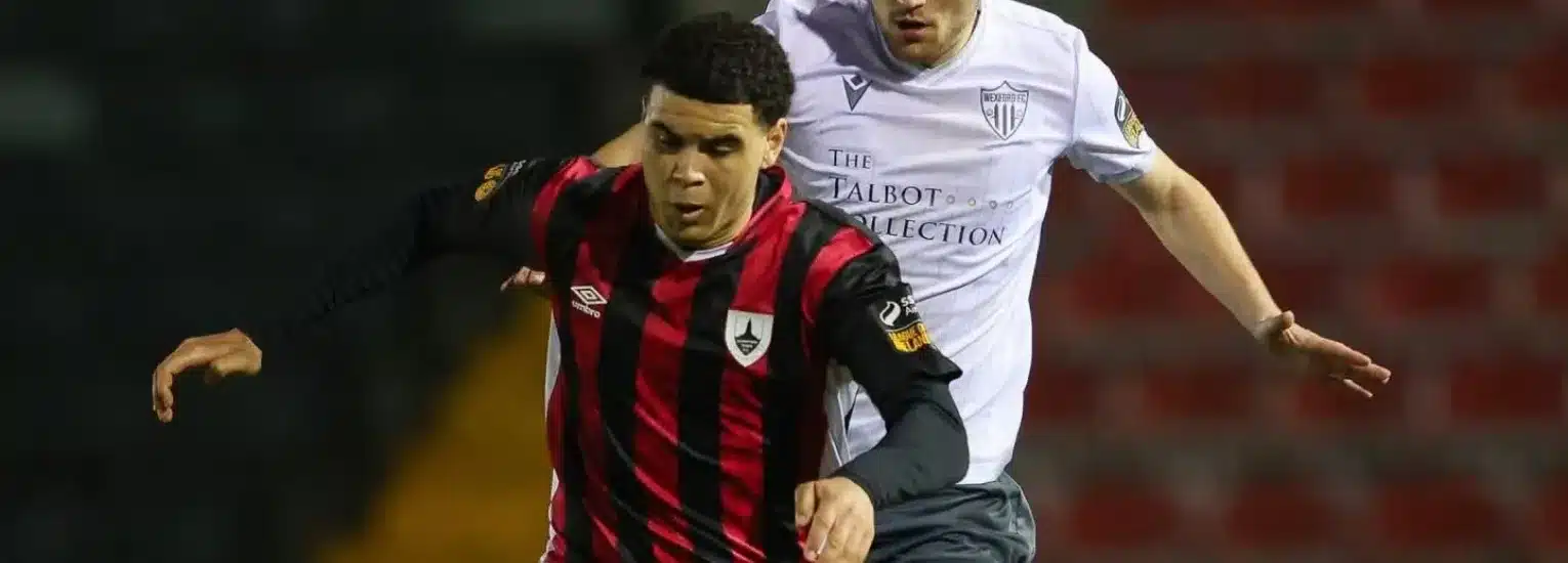 A footballer in a black and red striped shirt is dribbling the ball whilst being closely pursued by an opponent in a white shirt on a pitch during a match.